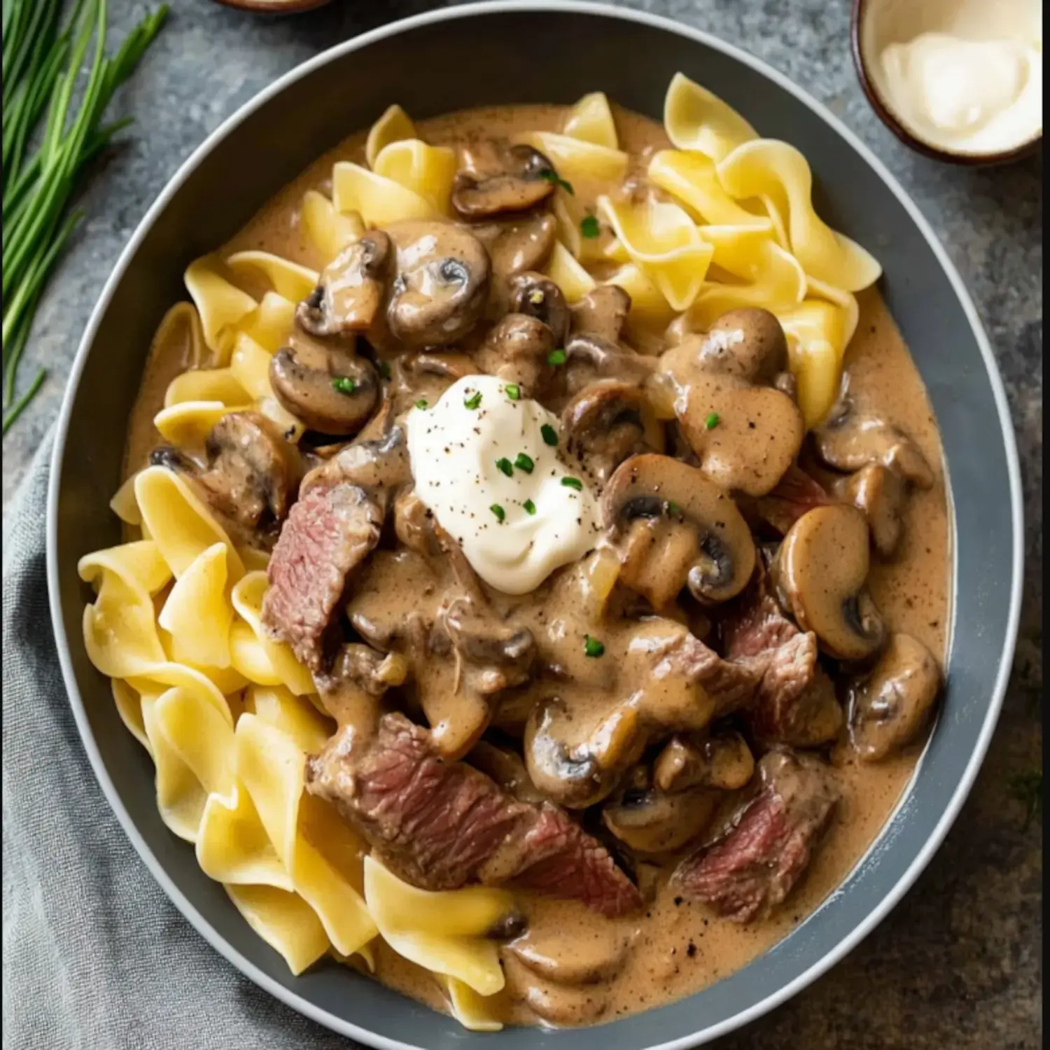 A bowl of egg noodles topped with beef, mushrooms, and cream sauce, garnished with a dollop of sour cream and chopped herbs.