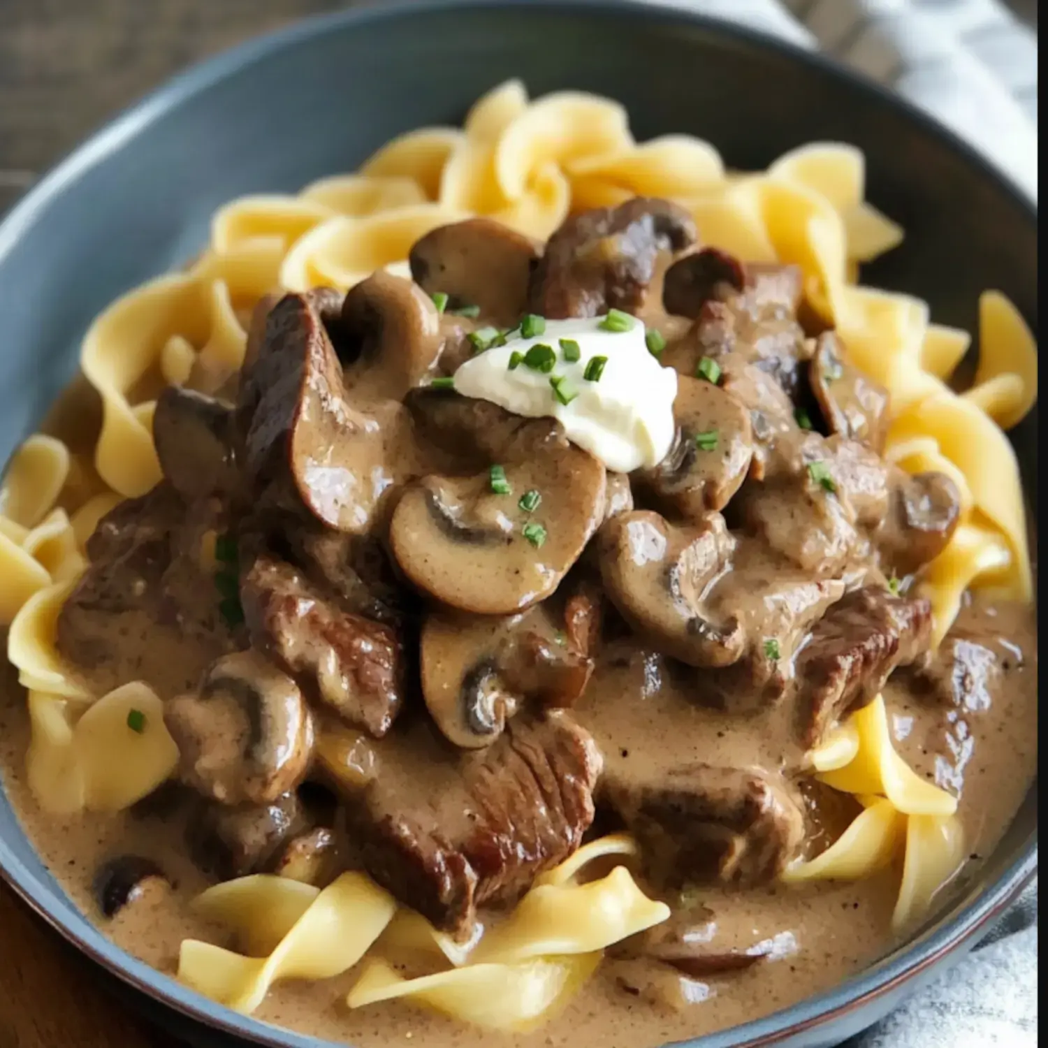 A serving of egg noodles topped with tender beef and mushrooms in a creamy sauce, garnished with a dollop of sour cream and chopped chives.