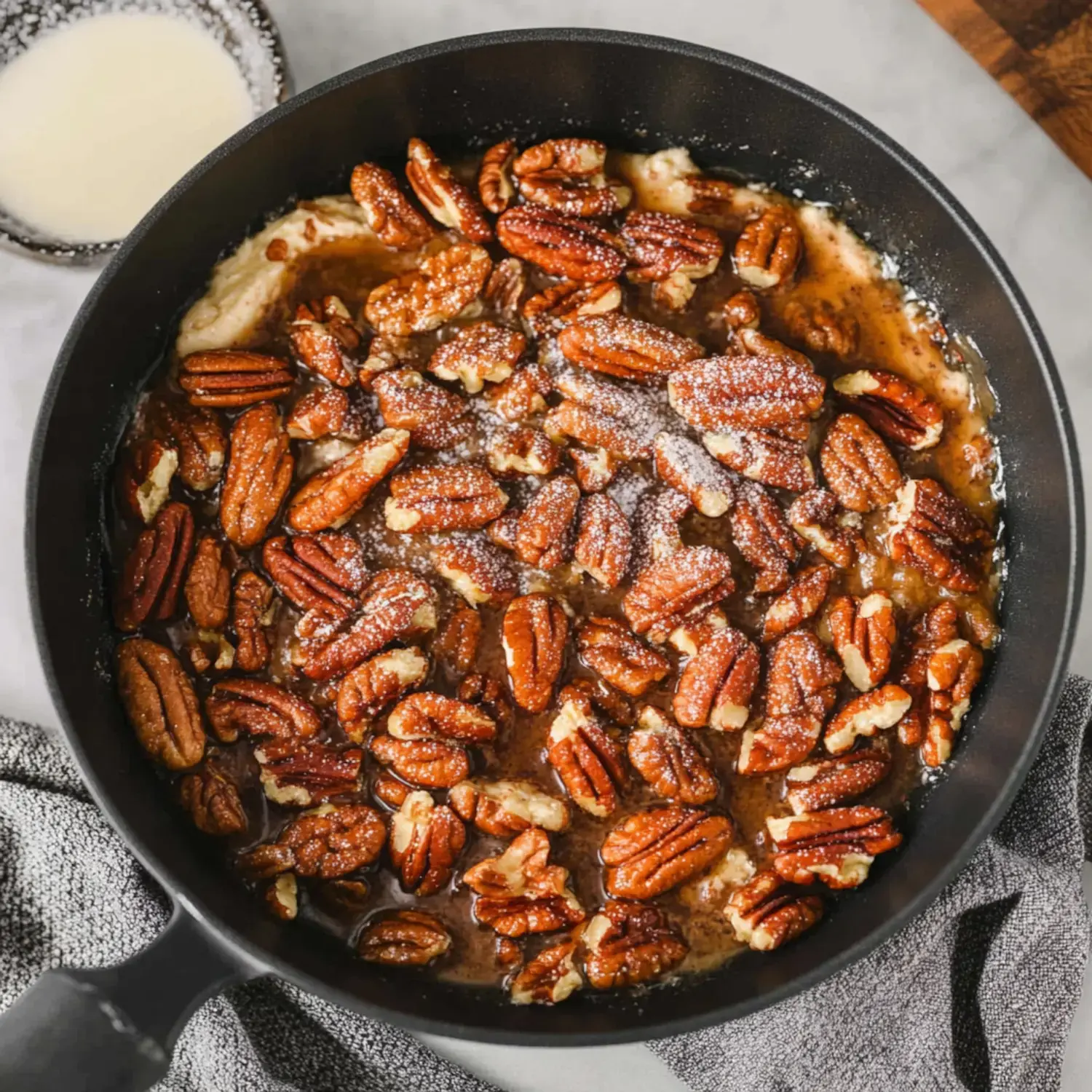 A close-up view of a skillet filled with glazed pecans, lightly dusted with powdered sugar, alongside a bowl of cream.