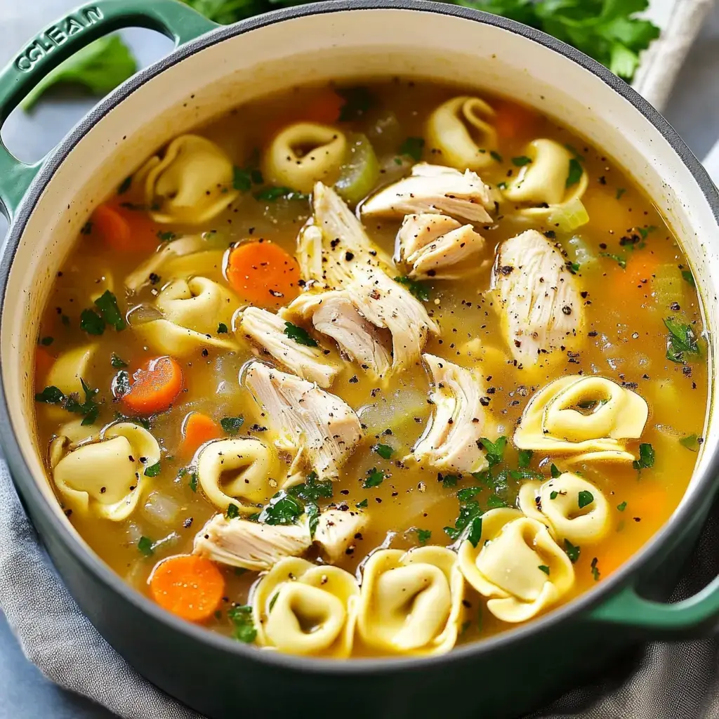 A close-up of a pot filled with chicken tortellini soup containing shredded chicken, carrots, and herbs.