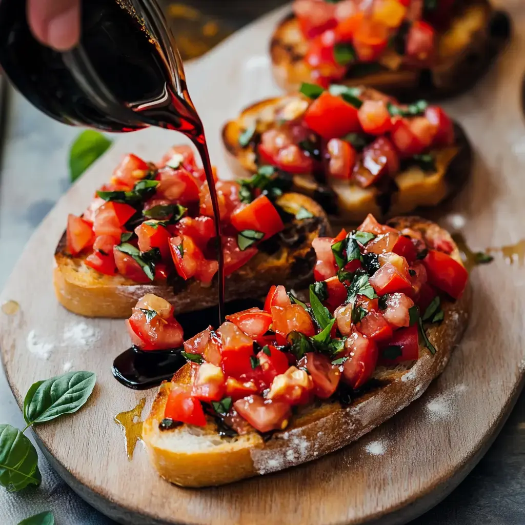 A hand is pouring balsamic glaze over bruschetta topped with diced tomatoes and fresh basil on a wooden serving board.