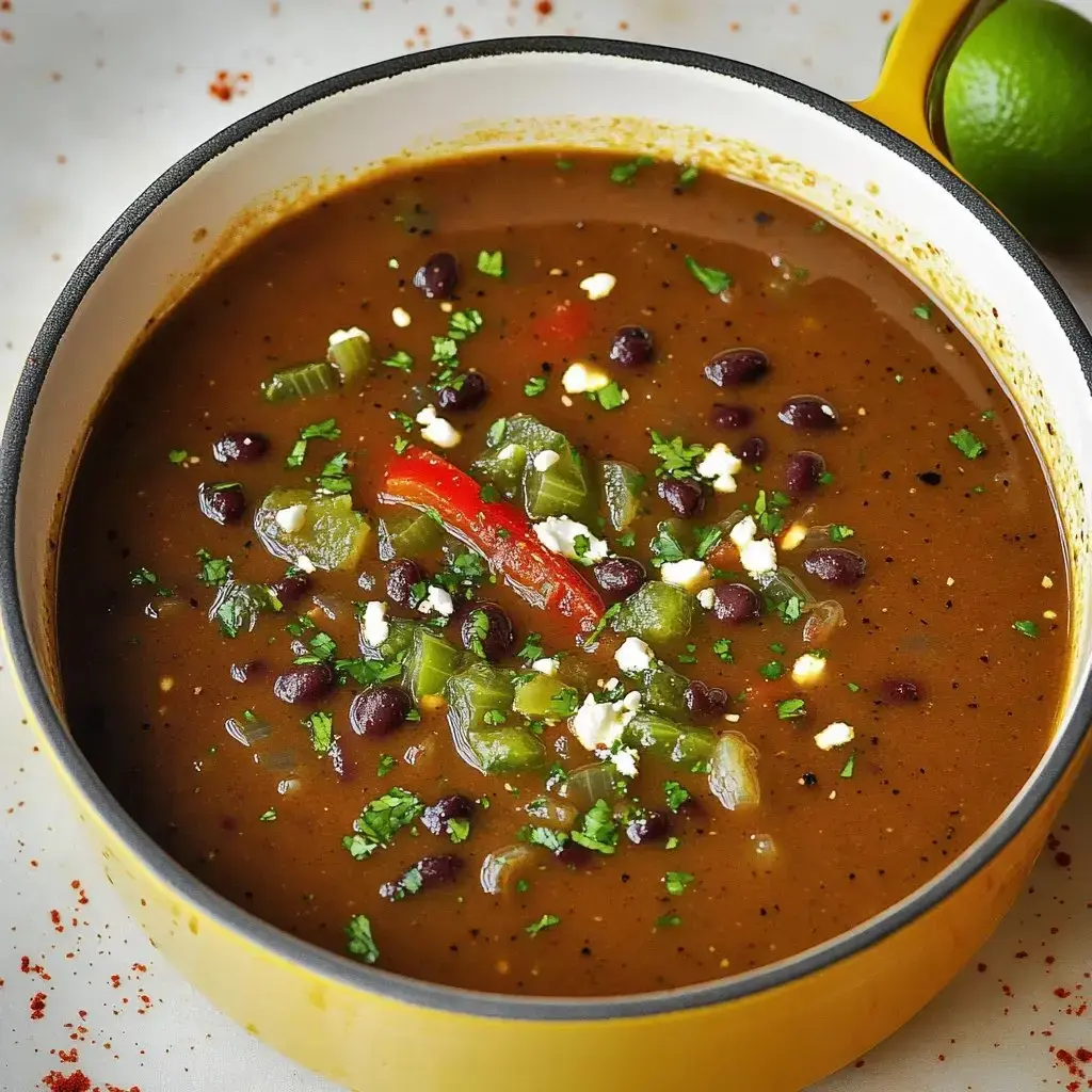 A vibrant bowl of black bean soup topped with chopped vegetables, herbs, and a slice of red pepper, with a lime in the background.