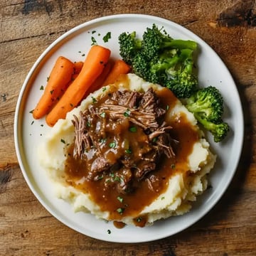 A plate of mashed potatoes topped with shredded beef in gravy, accompanied by steamed broccoli and carrots.